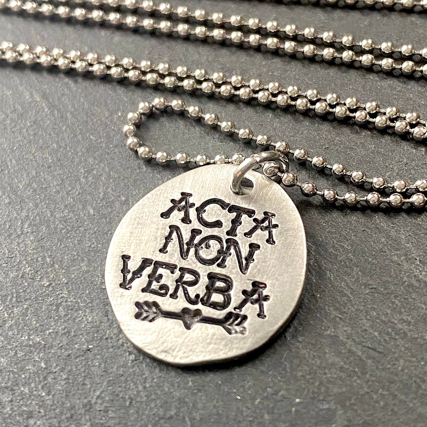 acta non verba latin phrase necklace. deeds not words hand stamped in tattoo font in pewter disc with heart arrow. drake designs jewelry